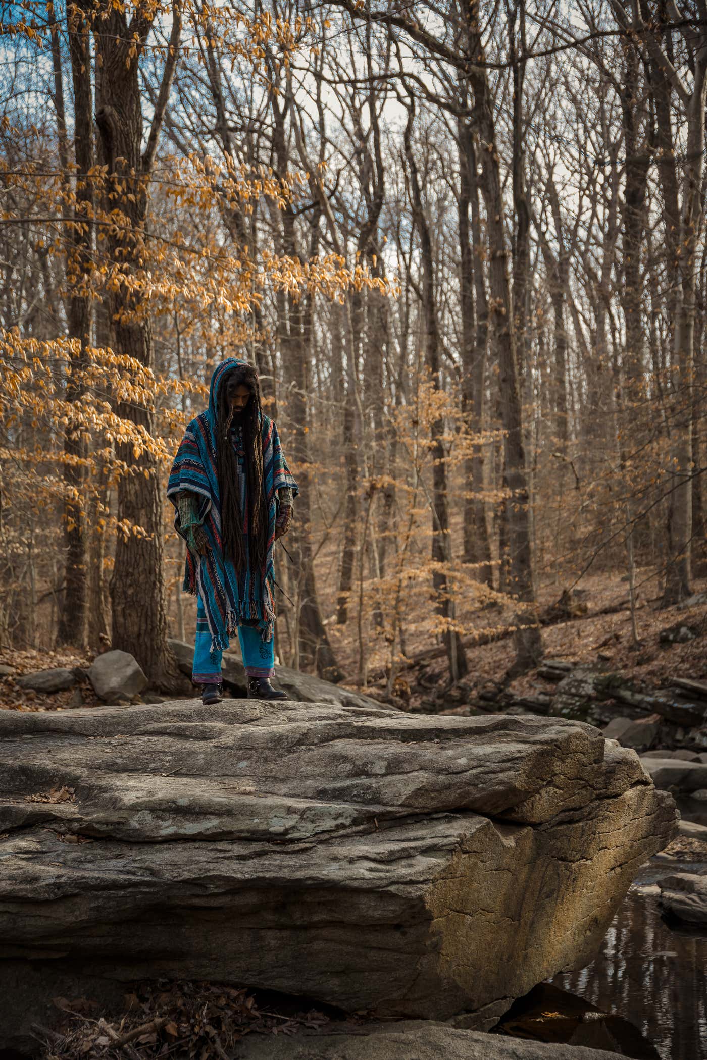 Model in a hooded striped Hippie Hut poncho standing on a rock ledge in bare winter woodland