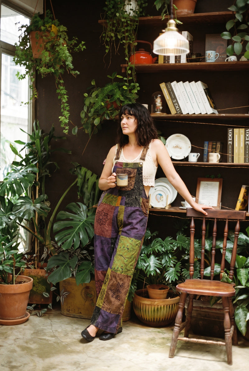Woman leaning on a shelf with a mug, wearing the Duskwood Overalls in aubergine, olive and rust patchwork with block-printed florals