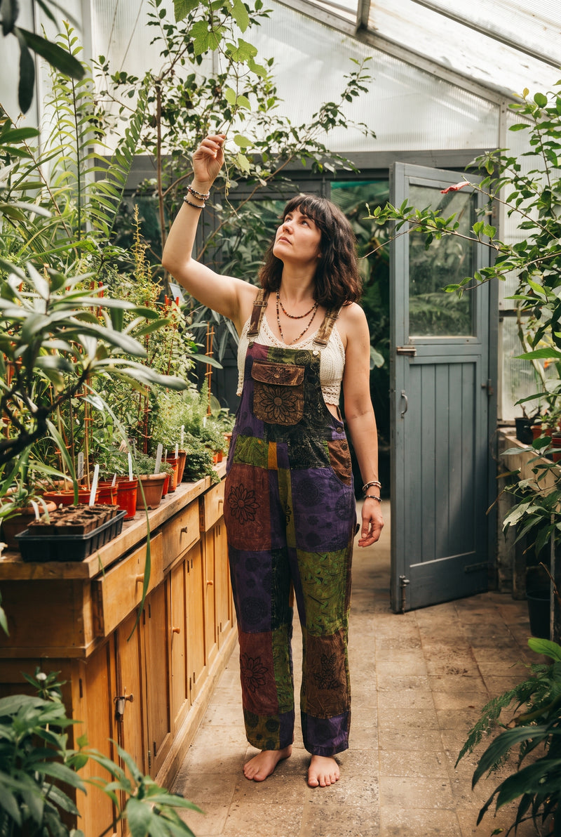 Woman reaching for a branch in a sunlit greenhouse, wearing the Duskwood Overalls in aubergine and olive patchwork over a crochet top