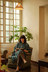 Tidewater Gheri Poncho worn seated in an armchair, teal stripe with a hood and fringed hem.