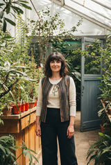 The Juniper Woven Vest in the greenhouse, braided tassel tie at the throat.