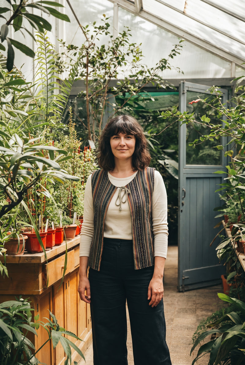 The Juniper Woven Vest in the greenhouse, braided tassel tie at the throat.