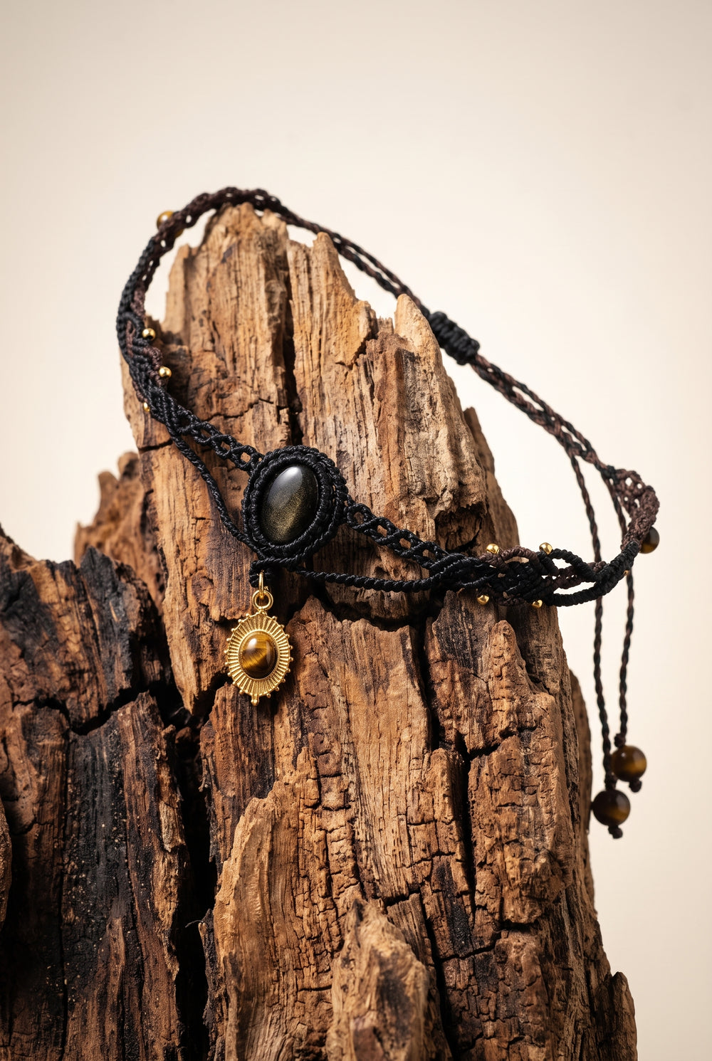 Obsidian Sunburst Choker with golden-sheen obsidian stone in black macramé cord and brass sunburst tiger's eye charm, draped over weathered driftwood.
