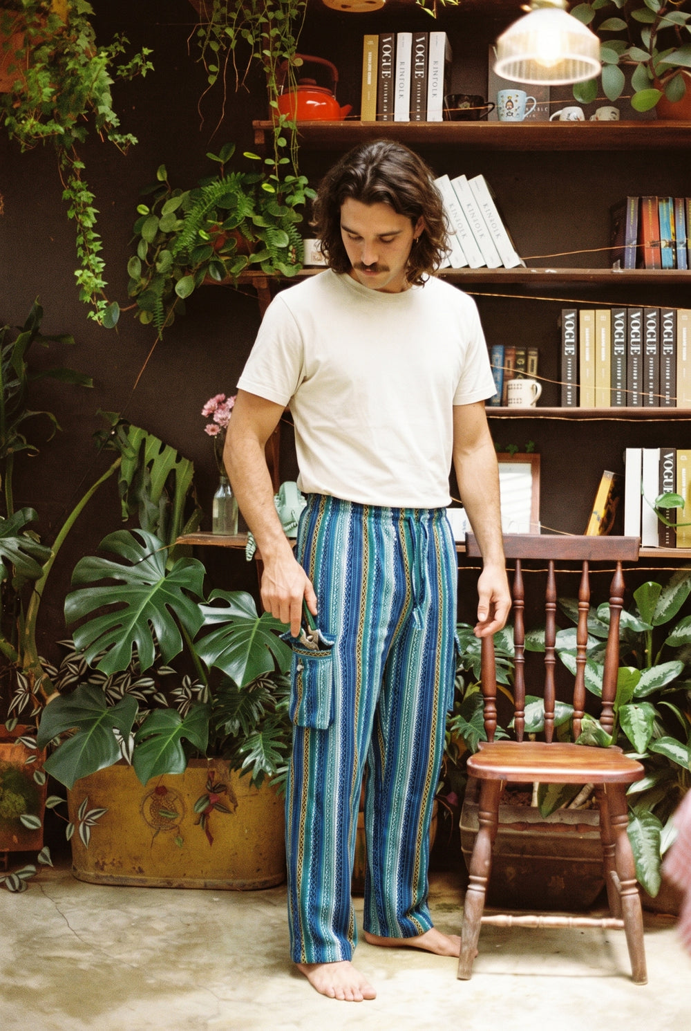 Man wearing the Riverstone Gheri Cargo Pants in blue and green woven stripe with a cream tee, reaching into the side cargo pocket