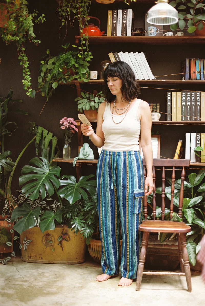 Woman in the Riverstone Gheri Cargo Pants and a cream singlet, standing among ferns and bookshelves