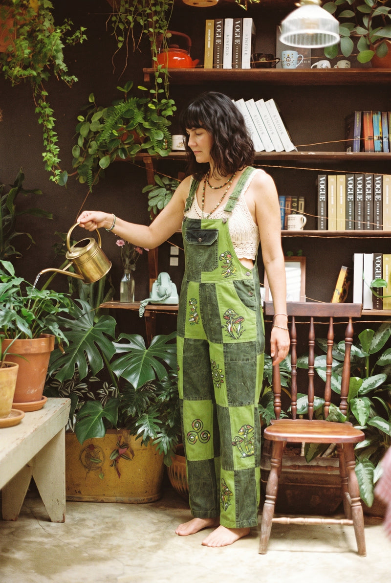 Woman watering plants in the Sungrove Overalls, lime and forest green checkerboard patchwork dungarees block-printed with mushrooms, over a cream crochet top