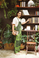 Man wearing the Sungrove Overalls, lime and forest green checkerboard patchwork dungarees, holding books in a plant-filled room