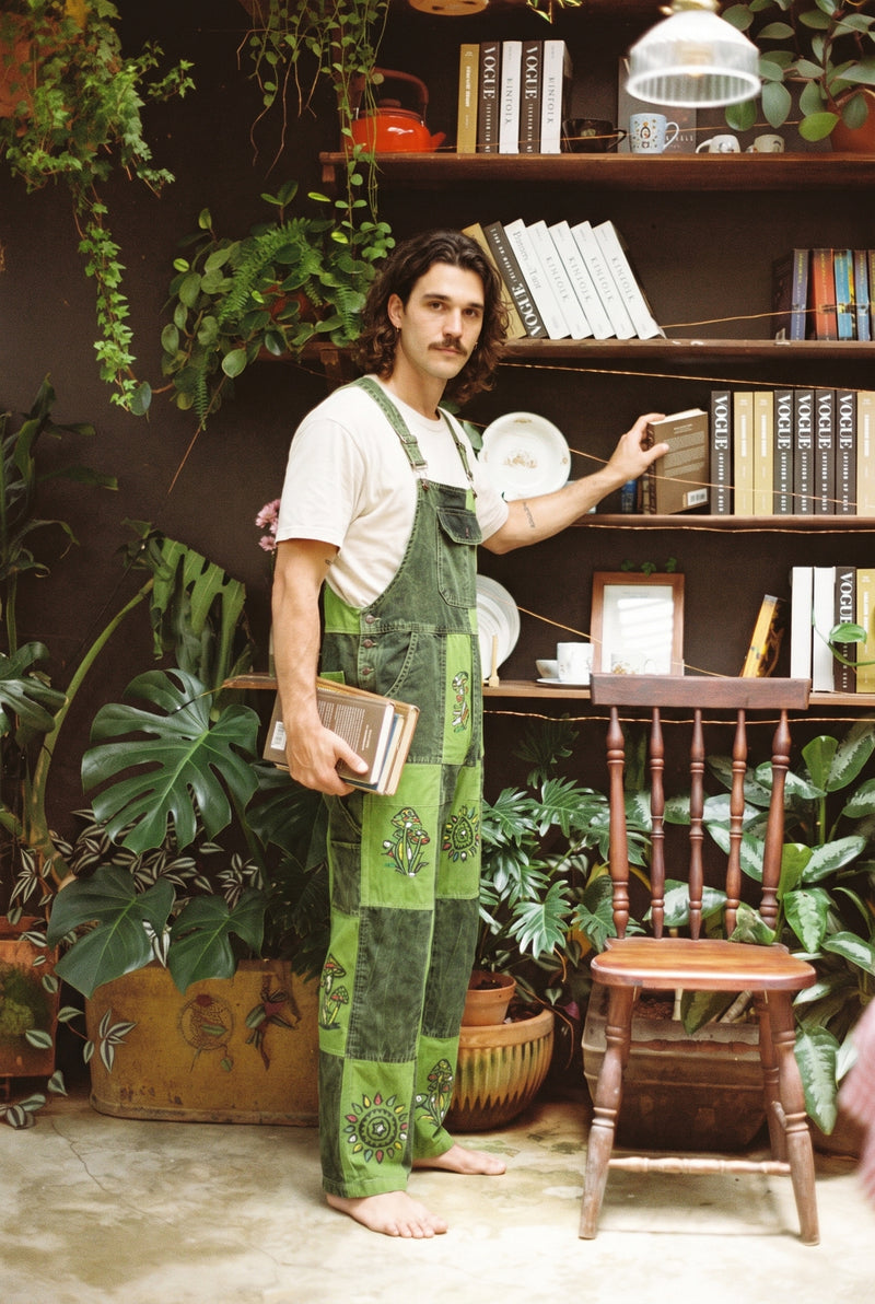 Man wearing the Sungrove Overalls, lime and forest green checkerboard patchwork dungarees, holding books in a plant-filled room