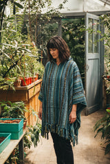 Woman in the Tidewater Gheri Poncho, teal and blue woven stripe with a fringed hem, among potted seedlings in a glasshouse