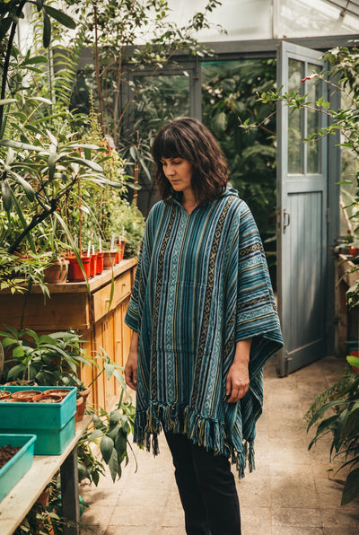 Woman in the Tidewater Gheri Poncho, teal and blue woven stripe with a fringed hem, among potted seedlings in a glasshouse #coll:womens-tops-outerwear #coll:boho-womens-clothing