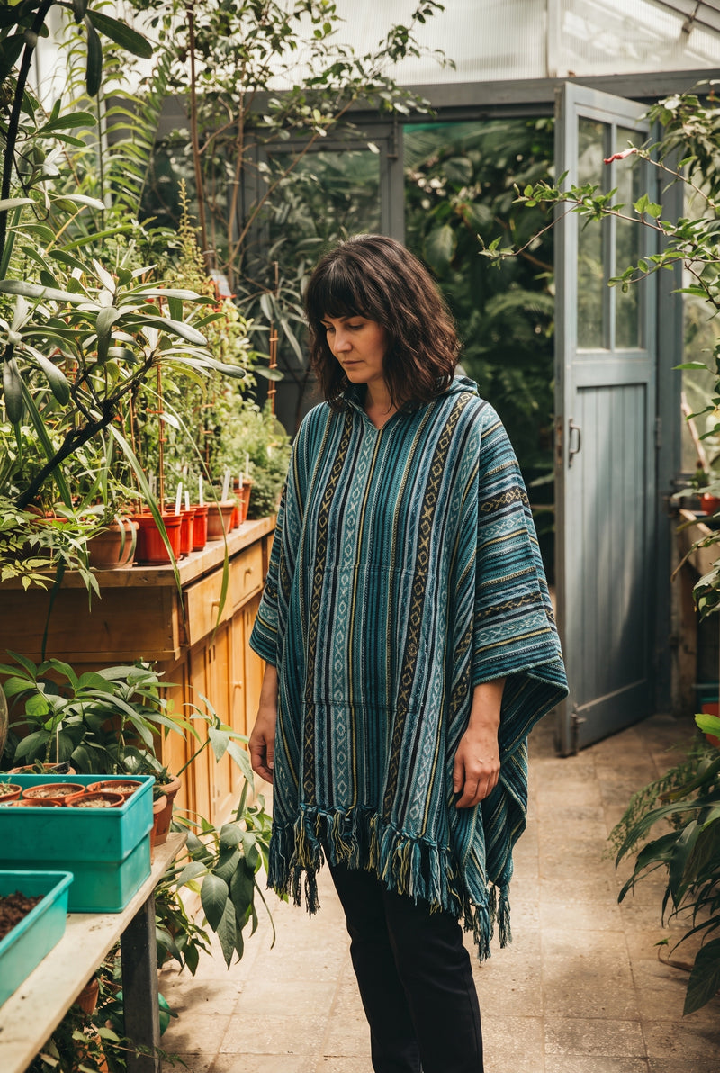 Woman in the Tidewater Gheri Poncho, teal and blue woven stripe with a fringed hem, among potted seedlings in a glasshouse
