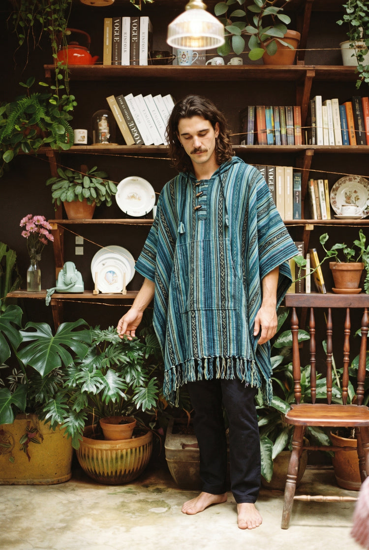 Man wearing the hooded Tidewater Gheri Poncho in teal stripe with toggle fastening, in front of a plant-filled bookshelf