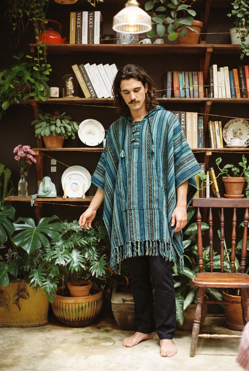 Man wearing the hooded Tidewater Gheri Poncho in teal stripe with toggle fastening, in front of a plant-filled bookshelf