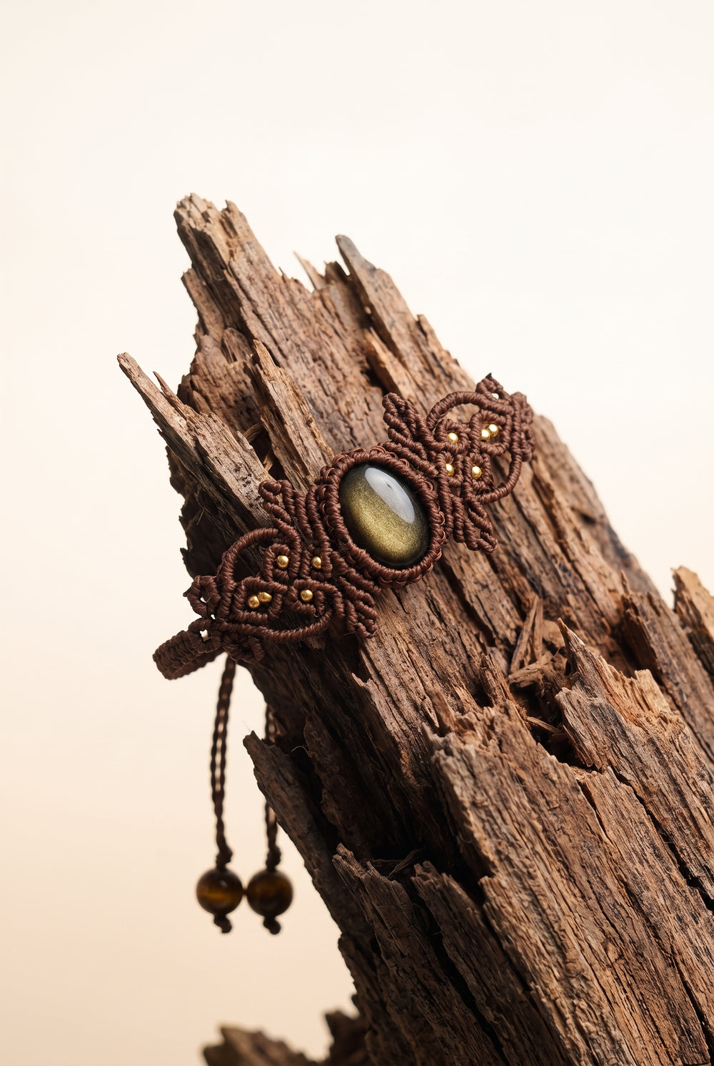 Woven Gold Obsidian Bracelet with golden-sheen obsidian stone in ornate brown macramé cord and brass beads, hanging on weathered driftwood.