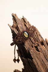 Woven Gold Obsidian Bracelet with golden-sheen obsidian stone in ornate brown macramé cord and brass beads, hanging on weathered driftwood.