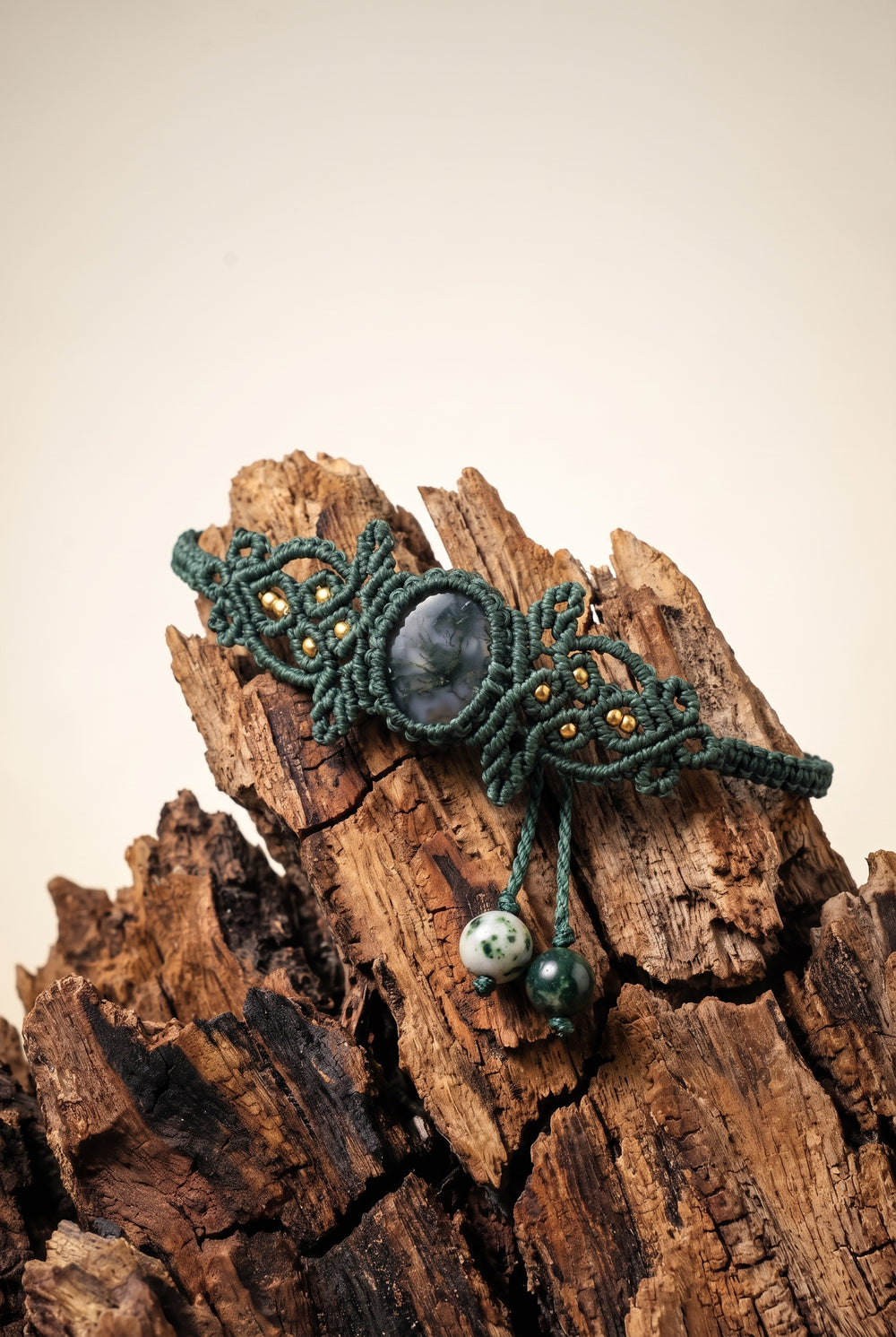 Woven Moss Agate Bracelet with dark moss agate stone in ornate green macramé cord and brass beads, draped over weathered driftwood.