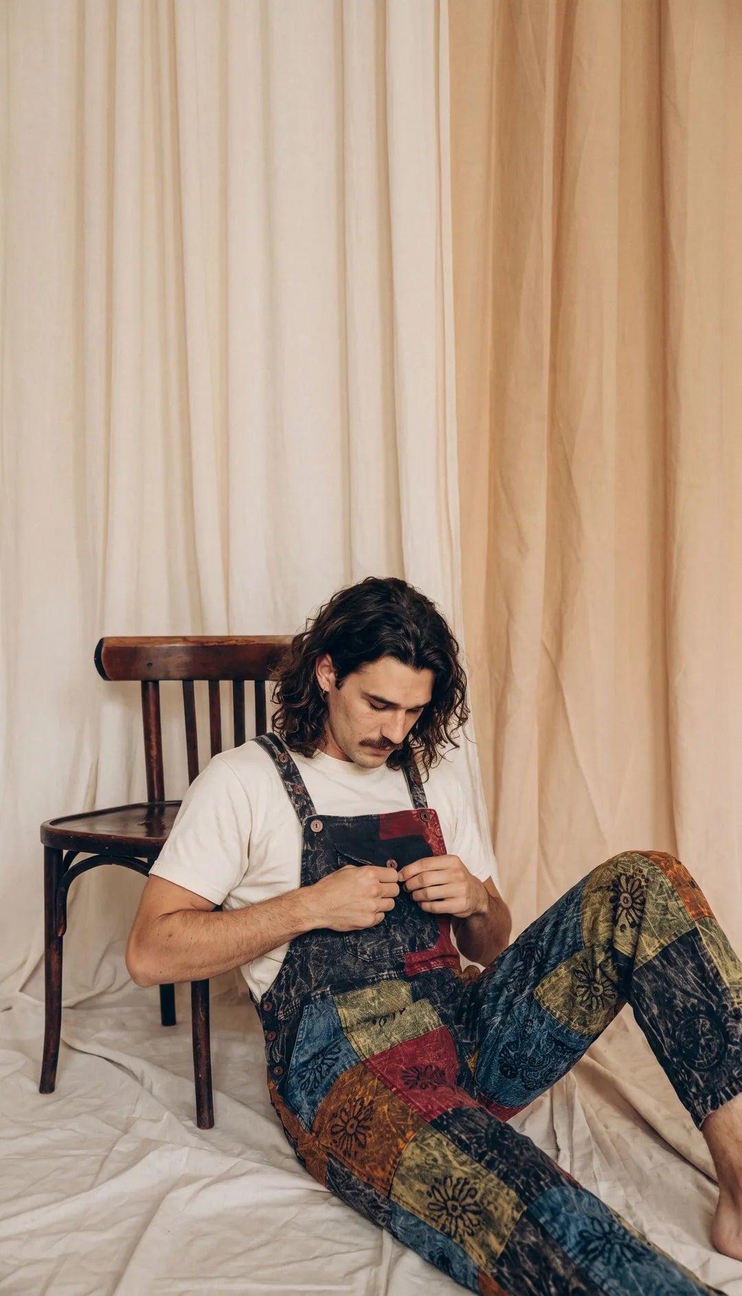A model in Emberfield Overalls sitting on the floor against a bentwood chair, calico drapes behind