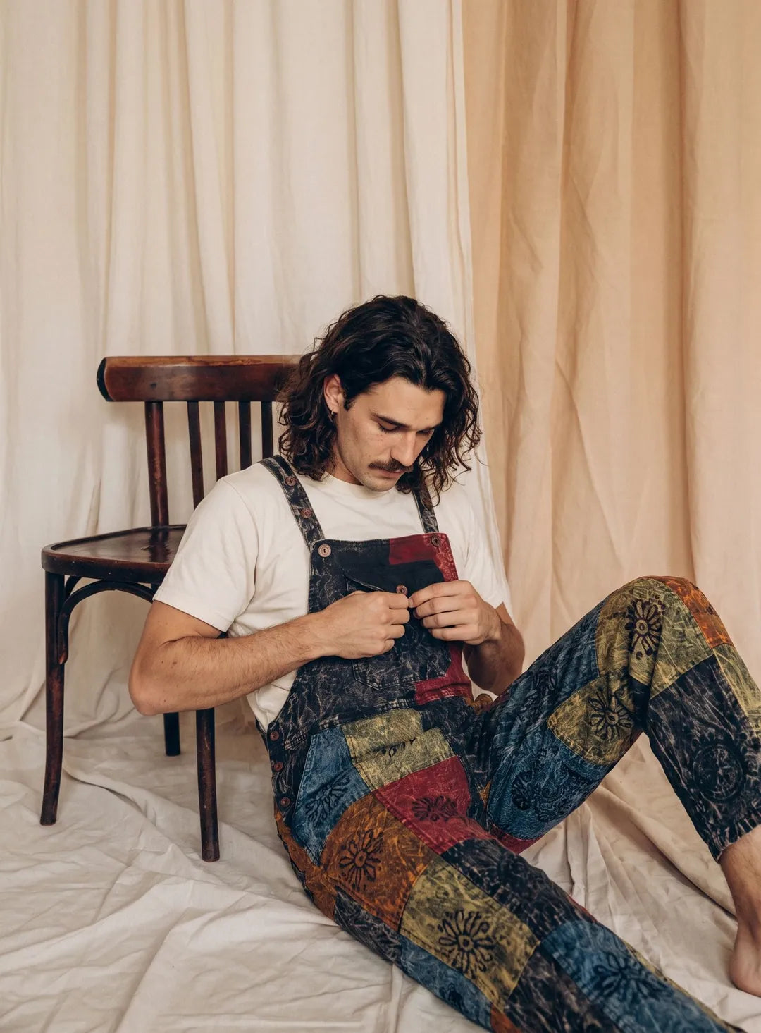 A model in Emberfield Overalls sitting on the floor against a bentwood chair, calico drapes behind