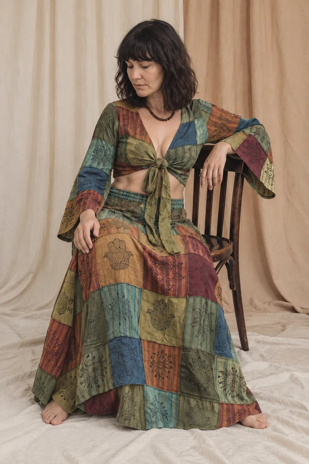 A model in the Fern and Fire Set, a patchwork tie-front top and maxi skirt in deep greens, ambers and reds, seated on a bentwood chair against calico drapes