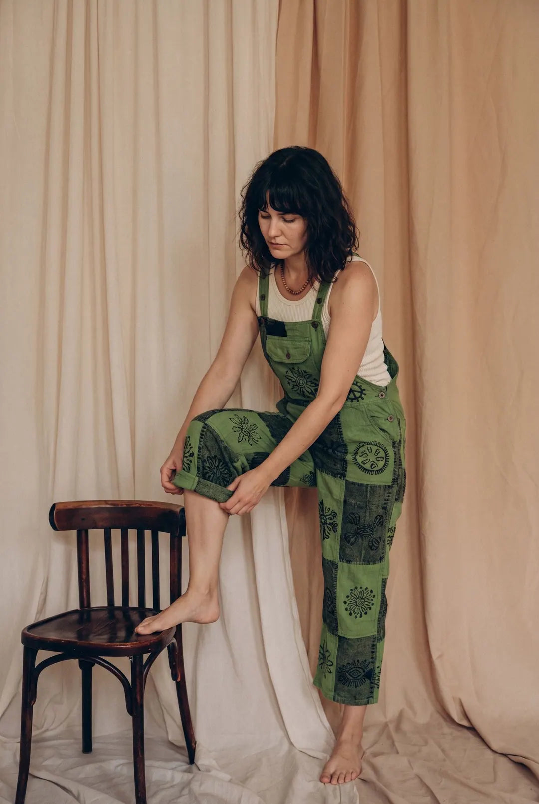 A model in Fernbrook Overalls, leaf and forest green patchwork, one foot on a bentwood chair against calico drapes