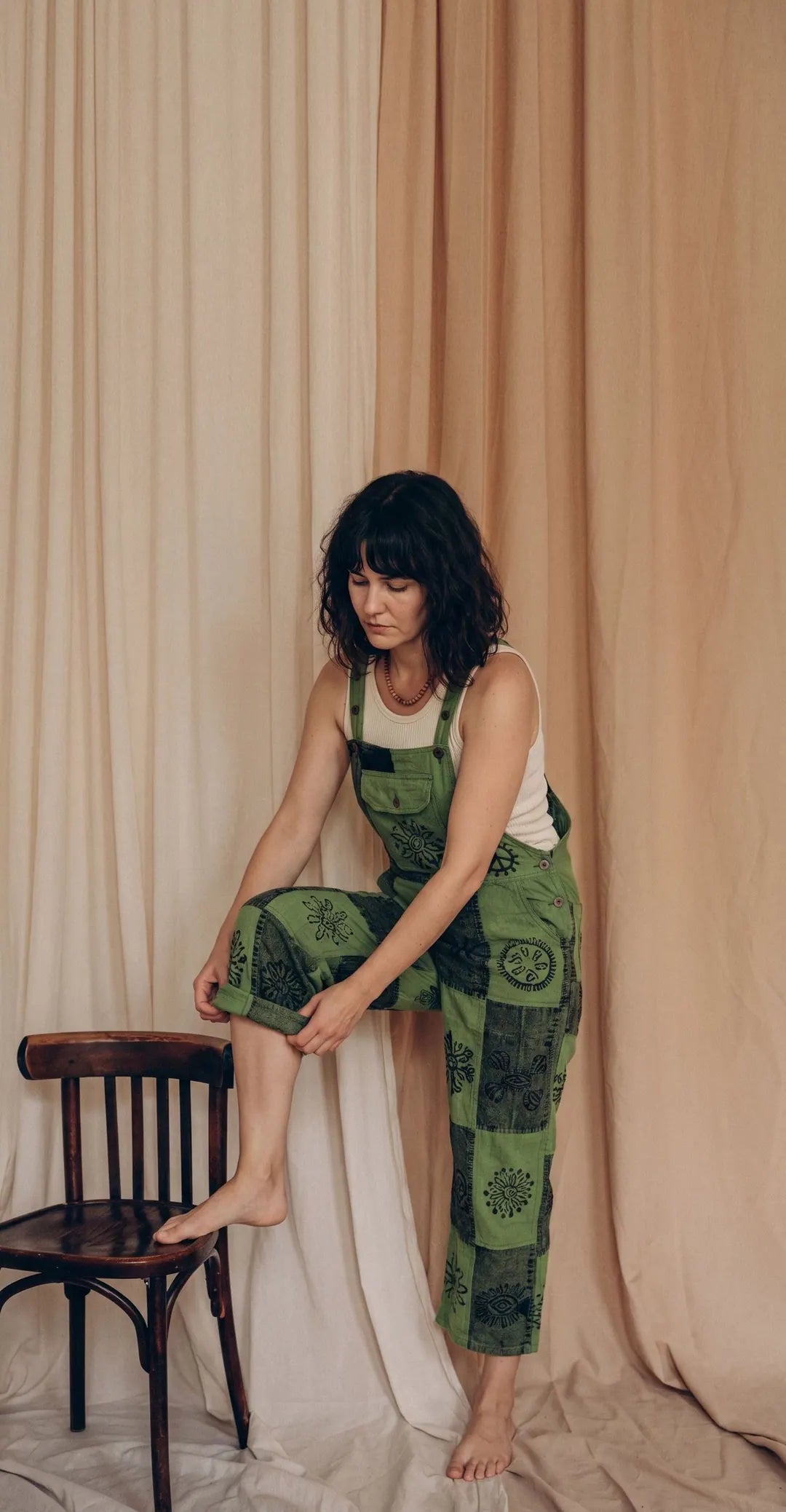 A model in Fernbrook Overalls, leaf and forest green patchwork, one foot on a bentwood chair against calico drapes