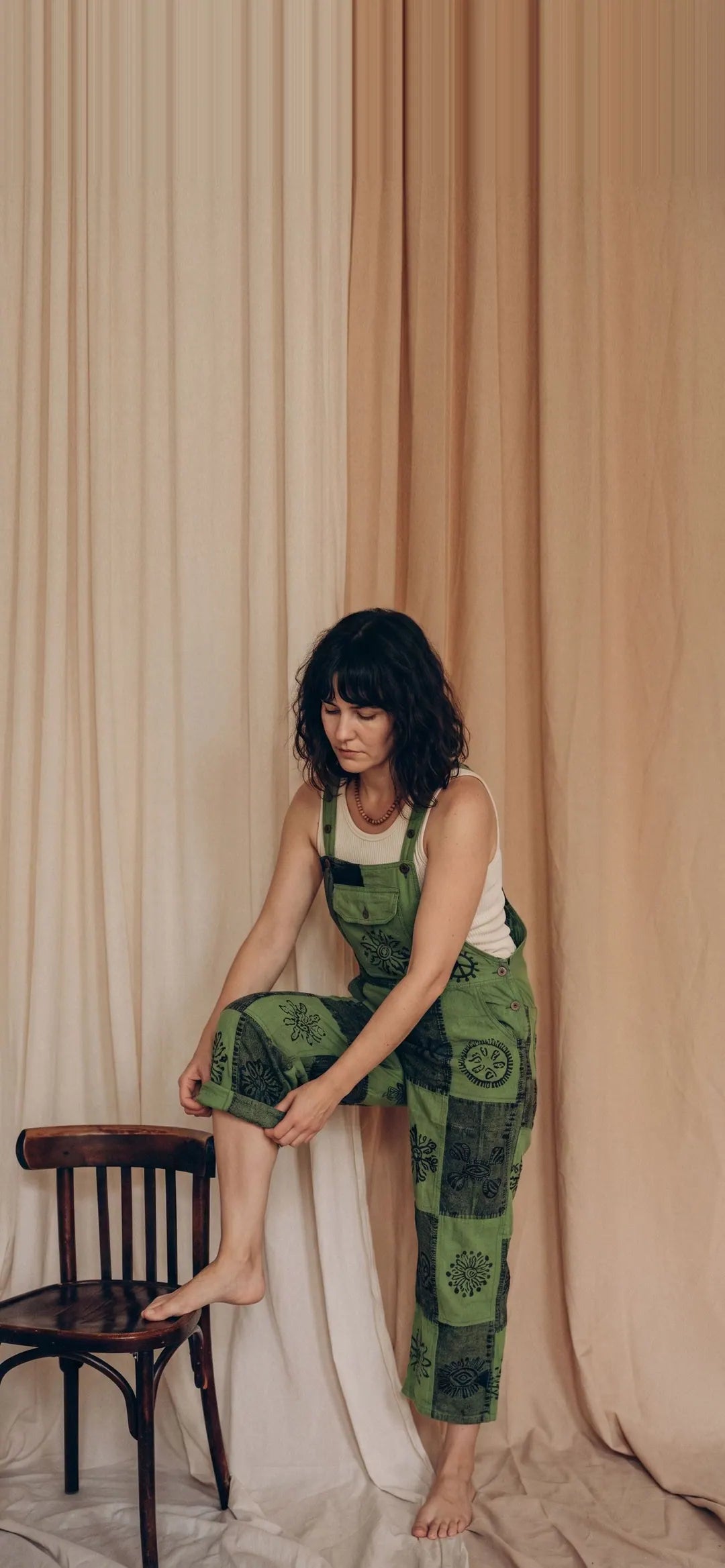 A model in Fernbrook Overalls, leaf and forest green patchwork, one foot on a bentwood chair against calico drapes
