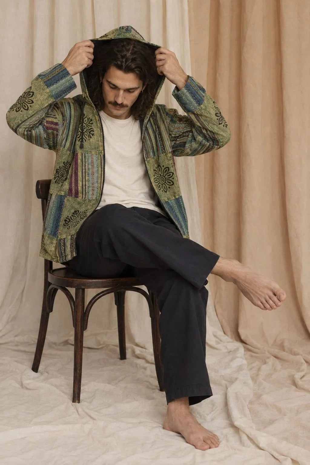 A model in the Green Horizon Patchwork Jacket, lime, brown, teal and rust patchwork, seated on a bentwood chair against calico drapes